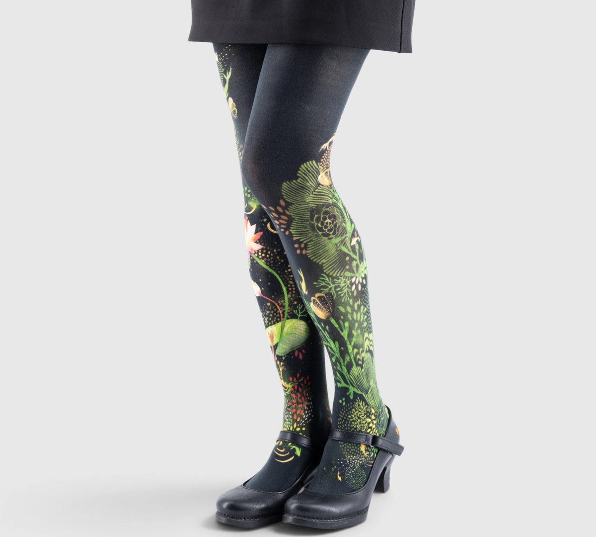 Jambes féminines en collants imprimés fleurs et plantes vertes sur fond noir, chaussures noires à talon moyen et robe sombre.